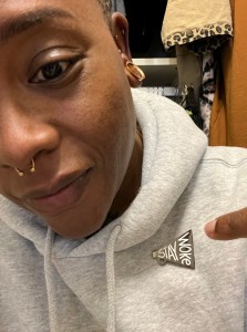 Black woman wearing gray hoodie sweatshirt pointing to a "Stay Woke" megaphone lapel pin