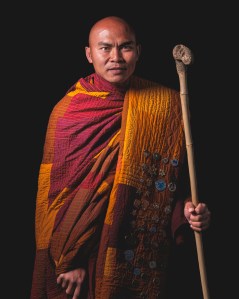 Bald head, brown skinned Monk wearing an orange and red robe and carrying a walking stick